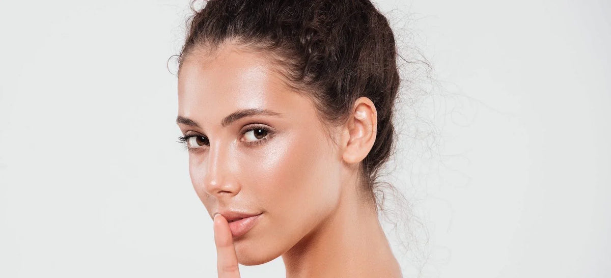 Woman with glowing, healthy skin holding a finger to her lips in a quiet gesture, showcasing a radiant complexion and smooth texture against a clean white background.