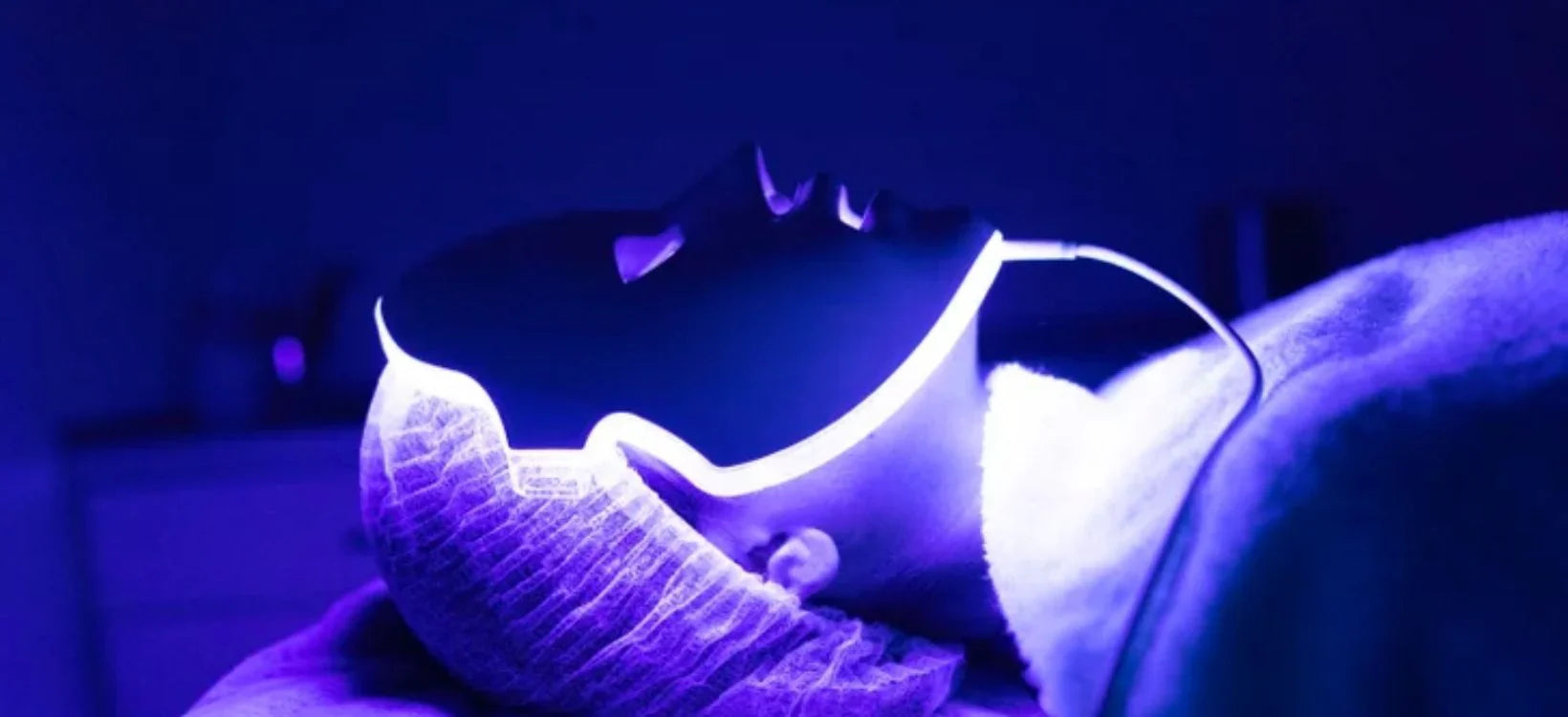 A person wearing an LED face mask emitting blue light, likely for skincare treatment, in a darkened spa or clinical setting.