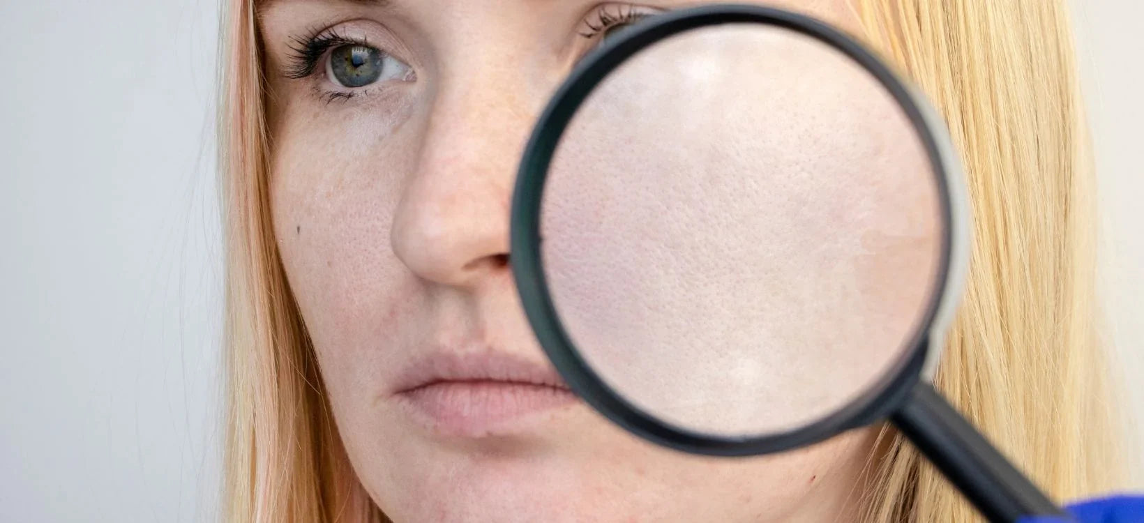 Close-up of a woman examining her skin pores with a magnifying glass.