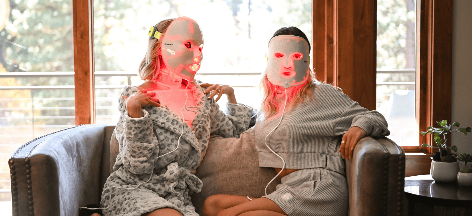 Two individuals wearing LED light therapy masks while relaxing on a couch, showcasing a red light treatment for skincare rejuvenation and anti-aging benefits.