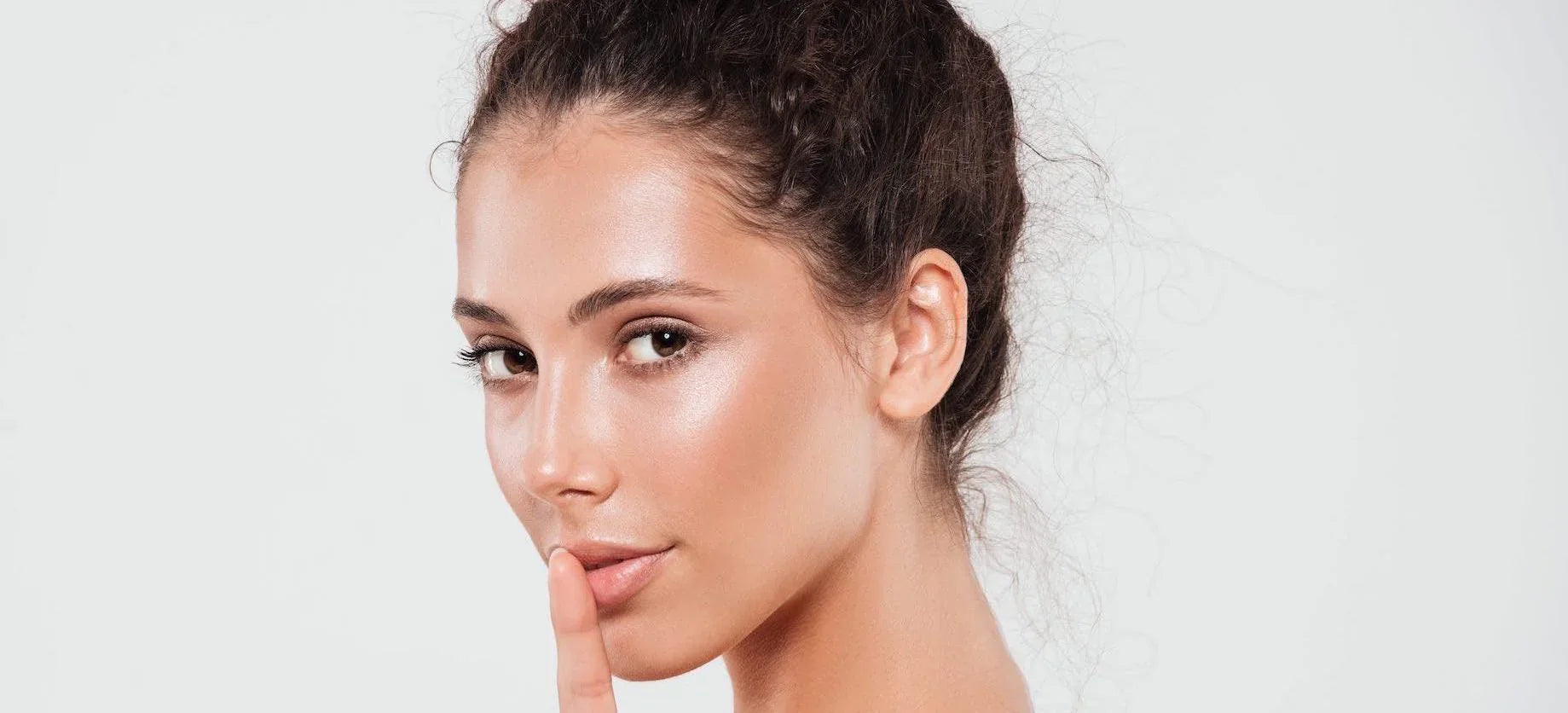 Close-up of a woman with glowing, clear skin and a natural makeup look, holding a finger to her lips, symbolizing a skincare secret or beauty tip.