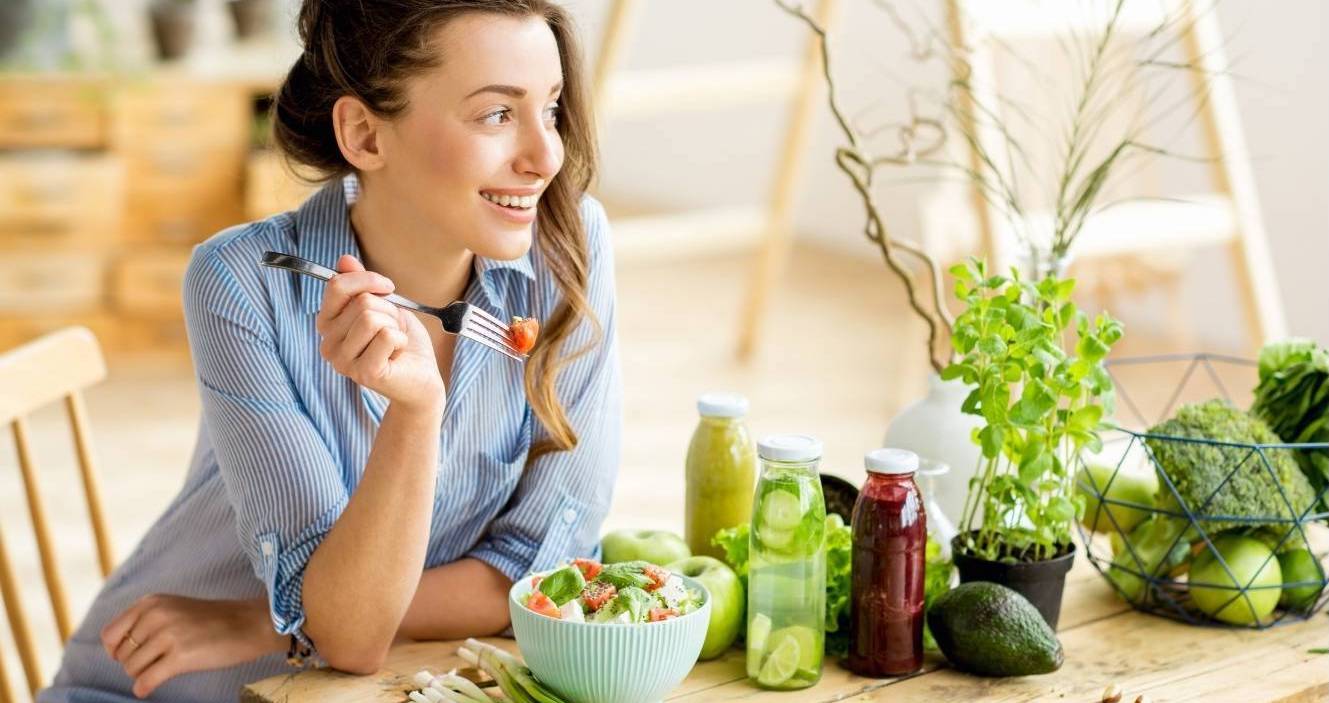 Woman enjoying a fresh, healthy salad with natural juice bottles, leafy greens, and fresh herbs on a wooden table, promoting a nutritious diet for wellness and anti-aging.