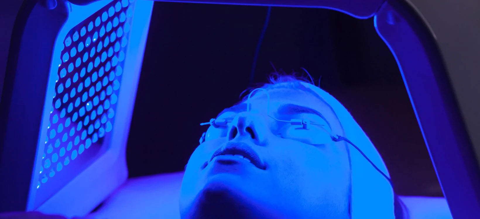 Person undergoing LED light therapy with protective eyewear, illuminated by a blue light treatment device for skincare benefits, in a dark setting.